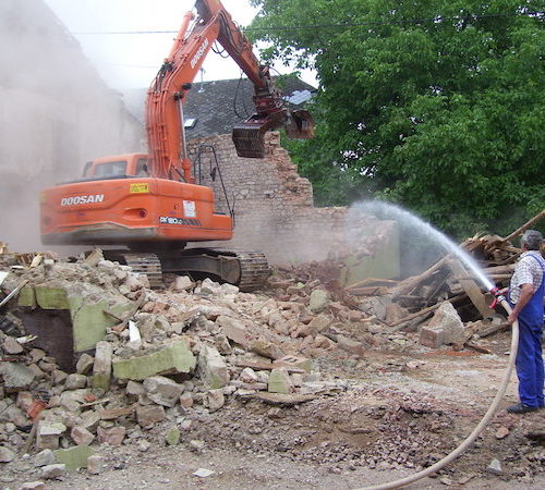 Ein Mann spritzt die Abbrucharbeiten eines Baggers an einer Mauer mit Wasser ab - Bauleistung